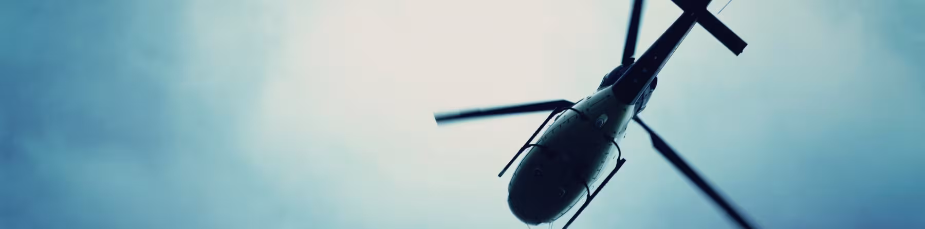 Helicopter seen from below in flight. Dark silhouette with spinning rotor blades against a cloudy blue sky.