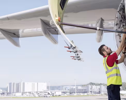 A SWISS line maintenance technician performing maintenance on an aircraft engine at the airport, with the engine panel open and airport facilities visible in the background.