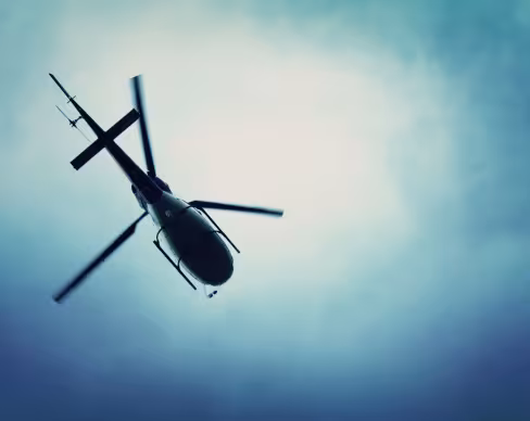 A helicopter viewed from below, flying against a cloudy sky with a blue gradient. The rotor blades appear slightly blurred, emphasizing motion.