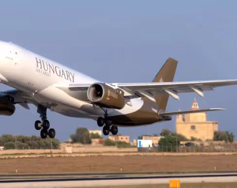 A Hungary-registered Airbus A330 takes off, with landing gear extended. The white fuselage features "HUNGARY" and gold-brown accents. The Hungarian flag colors appear on the winglets, with a historical building in the background.