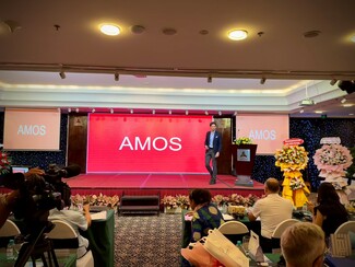 A man stands on a stage with a red screen behind him displaying the word "AMOS." People are seated at tables in front of the stage, with flower arrangements on either side.