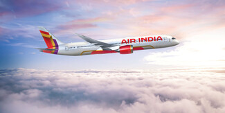 Air India airplane in flight, cruising over a sea of white clouds with a serene sky tinged in pink and blue hues. The aircraft’s red-and-orange tail and engine accents stand out against the pale sky.