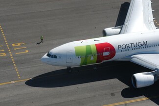 Top-down view of a TAP Air Portugal plane with a red and green logo, taxiing on the runway. A worker in a reflective vest is visible on the left, walking along the concrete near the aircraft.